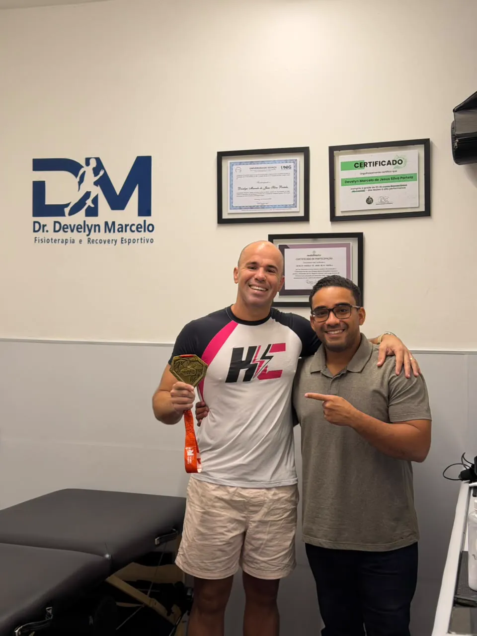 Dr. Develyn e paciente exibindo medalha no consultório, com a logo DM na parede