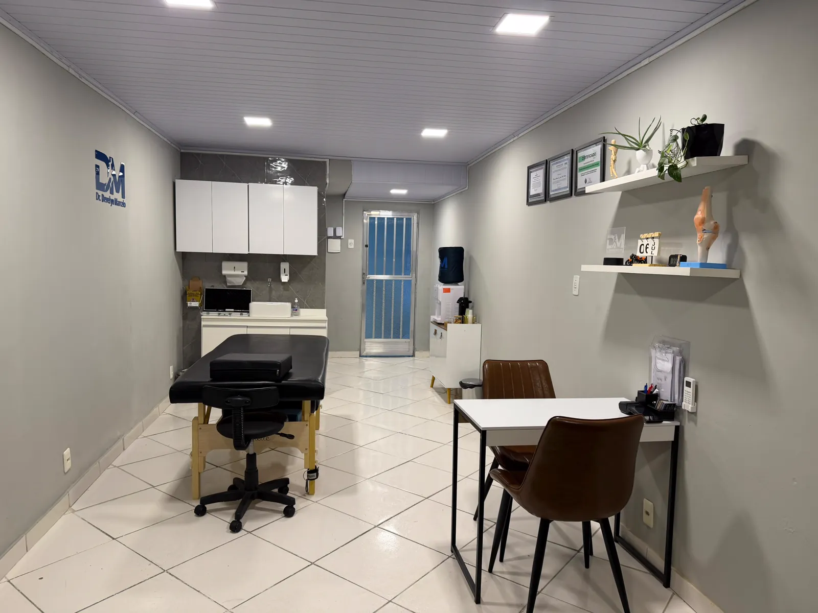 Sala de atendimento da DM: maca, banqueta, armários e pia, bebedouro e prateleiras com certificados e modelo anatômico do joelho