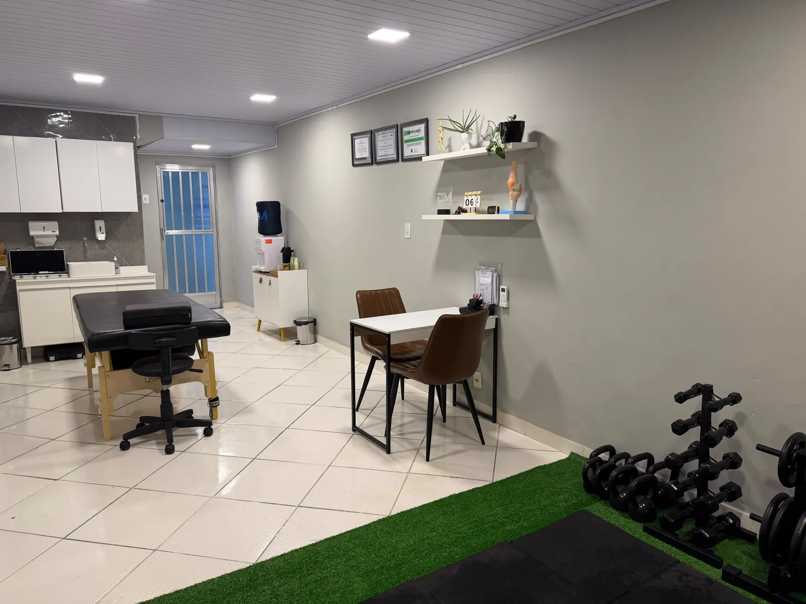 Sala de atendimento em outro ângulo, com mesa de avaliação, prateleiras com certificados e rack de halteres sobre grama sintética