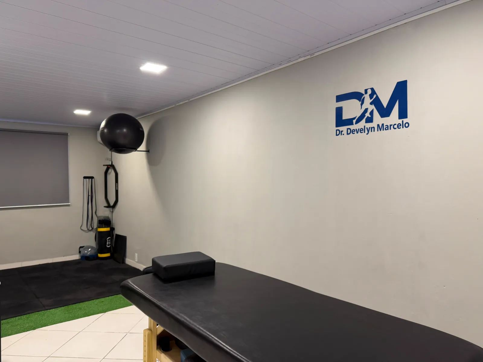 Maca de atendimento com a logo Dr. Develyn Marcelo na parede e a área de exercícios ao fundo