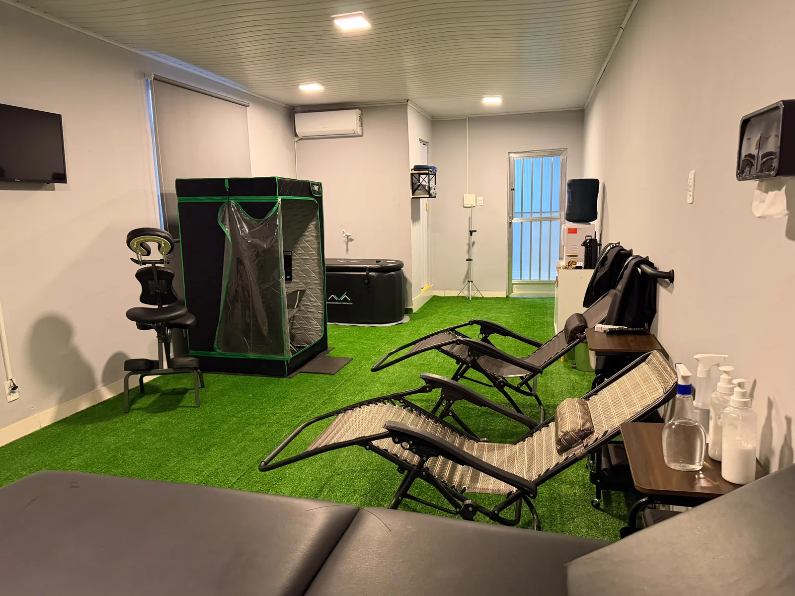 Sala de recovery com sauna infravermelho, banheira de imersão para crioterapia e cadeiras zero-gravity sobre grama sintética